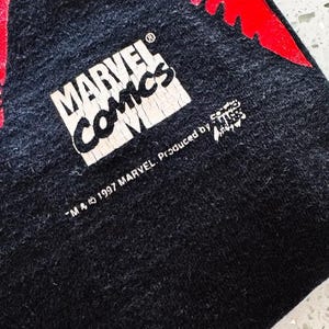 May include: A black fabric garment featuring a white Marvel Comics logo with visible cracking in the print. Below the logo, the text reads TM & 1997 MARVEL. Produced by and includes a small graphic element. A portion of a red design is visible.