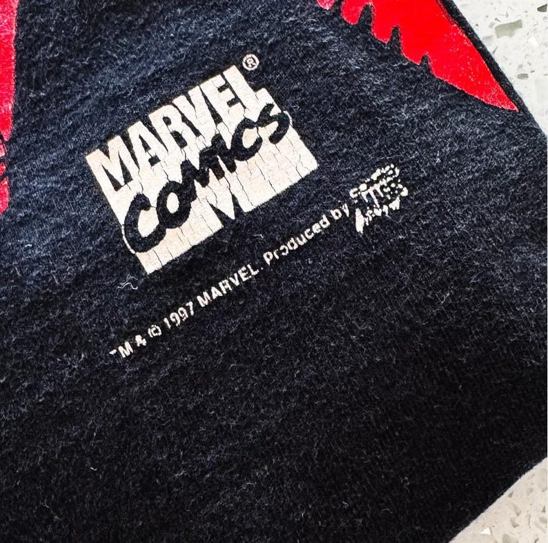May include: A black fabric garment featuring a white Marvel Comics logo with visible cracking in the print. Below the logo, the text reads TM & 1997 MARVEL. Produced by and includes a small graphic element. A portion of a red design is visible.