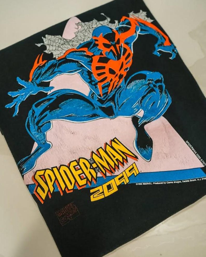 Op de afbeelding: Een zwart t-shirt met een grafische print van Spider-Man 2099 in blauw en oranje. Het personage staat in een dynamische pose boven de gele en oranje tekst die SPIDER-MAN 2099 leest. De print bevat de Marvel copyright-tekst.
