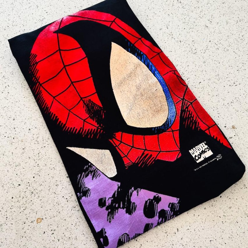 May include: A black t-shirt featuring a large graphic of the Spider-Man mask in red with black webbing details and white eye lenses. The design includes a purple abstract shape at the bottom and a white Marvel Comics logo in the lower right corner.