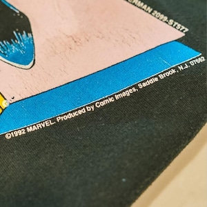 Op de afbeelding: Een zwart stoffen oppervlak met een grafische print met een roze en blauwe rand. De tekst luidt SPIDERMAN 2099-ST577 en 1992 MARVEL. Produced by Comic Images, Saddle Brook, N.J. 07662. De print bevat een gedeeltelijke blauwe en zwarte illustratie.