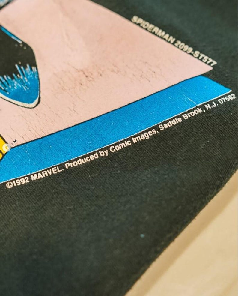 Op de afbeelding: Een zwart stoffen oppervlak met een grafische print met een roze en blauwe rand. De tekst luidt SPIDERMAN 2099-ST577 en 1992 MARVEL. Produced by Comic Images, Saddle Brook, N.J. 07662. De print bevat een gedeeltelijke blauwe en zwarte illustratie.