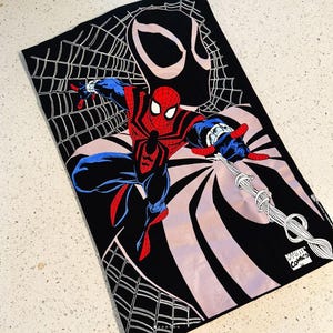 May include: A black rectangular towel featuring a graphic print of Spider-Man in a red and blue suit, positioned in front of a large black mask silhouette and a white spider web pattern. The bottom corner includes the Marvel Comics logo text.