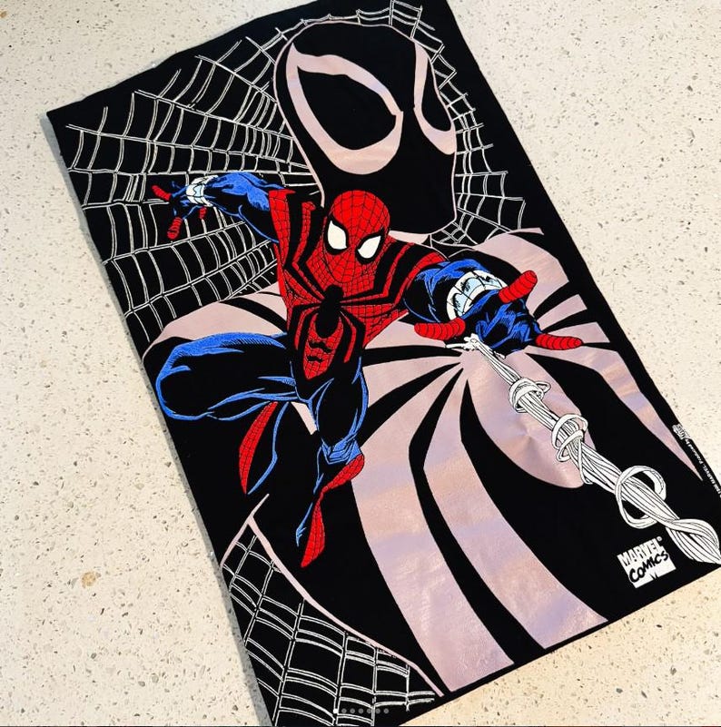 May include: A black rectangular towel featuring a graphic print of Spider-Man in a red and blue suit, positioned in front of a large black mask silhouette and a white spider web pattern. The bottom corner includes the Marvel Comics logo text.