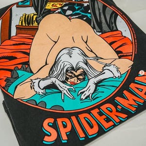 May include: A black t-shirt featuring a circular graphic of a comic book character with long white hair, a black mask, and white gloves, lying on a bed with orange sheets and a teal blanket. The text SPIDER-MAN is printed in orange below the circle.