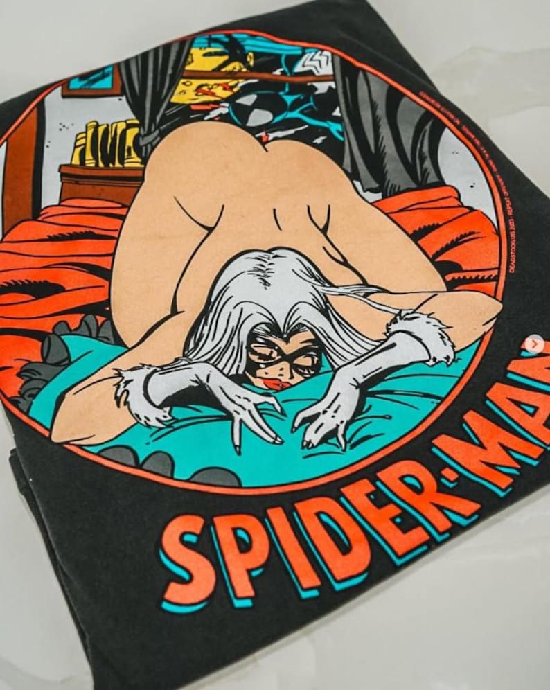 May include: A black t-shirt featuring a circular graphic of a comic book character with long white hair, a black mask, and white gloves, lying on a bed with orange sheets and a teal blanket. The text SPIDER-MAN is printed in orange below the circle.
