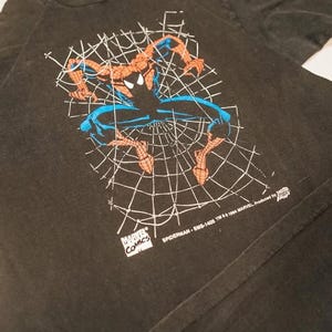 May include: A black short-sleeved t-shirt featuring a graphic of Spider-Man in a crouching position against a white spiderweb background. The print includes the text Marvel Comics, Spiderman-SMS-1405 TM & 1994 Marvel, and a small logo.