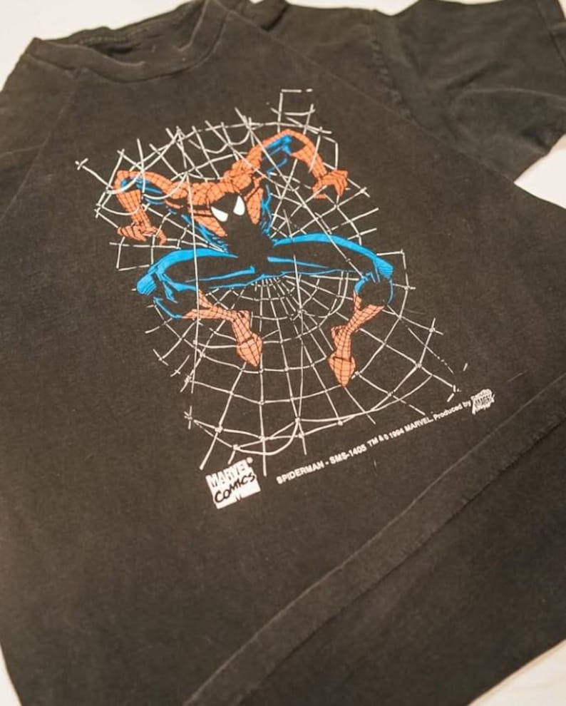 May include: A black short-sleeved t-shirt featuring a graphic of Spider-Man in a crouching position against a white spiderweb background. The print includes the text Marvel Comics, Spiderman-SMS-1405 TM & 1994 Marvel, and a small logo.