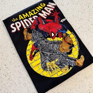K&ouml;nnte beinhalten: Ein schwarzes T-Shirt mit einer Grafik von The Amazing Spider-Man in Rot und Blau, die eine in Spinnweben eingewickelte Figur vor einem gelben, kreisf&ouml;rmigen Ziegelhintergrund zeigt. Der Text lautet The Amazing Spider-Man und enth&auml;lt eine Sprechblase.