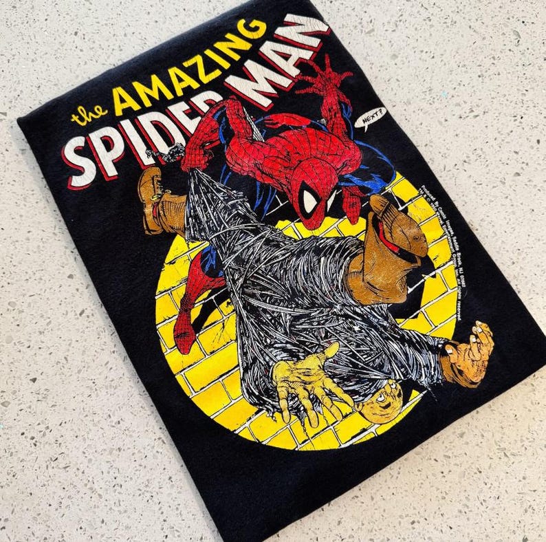 K&ouml;nnte beinhalten: Ein schwarzes T-Shirt mit einer Grafik von The Amazing Spider-Man in Rot und Blau, die eine in Spinnweben eingewickelte Figur vor einem gelben, kreisf&ouml;rmigen Ziegelhintergrund zeigt. Der Text lautet The Amazing Spider-Man und enth&auml;lt eine Sprechblase.