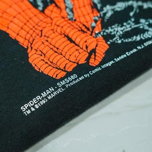 May include: A black fabric garment featuring a bright orange graphic of a gloved hand with a webbed pattern. The text reads: SPIDER-MAN - SMS680 TM & &copy;1993 MARVEL. Produced by Comic Images, Saddle Brook, N.J. 07662.