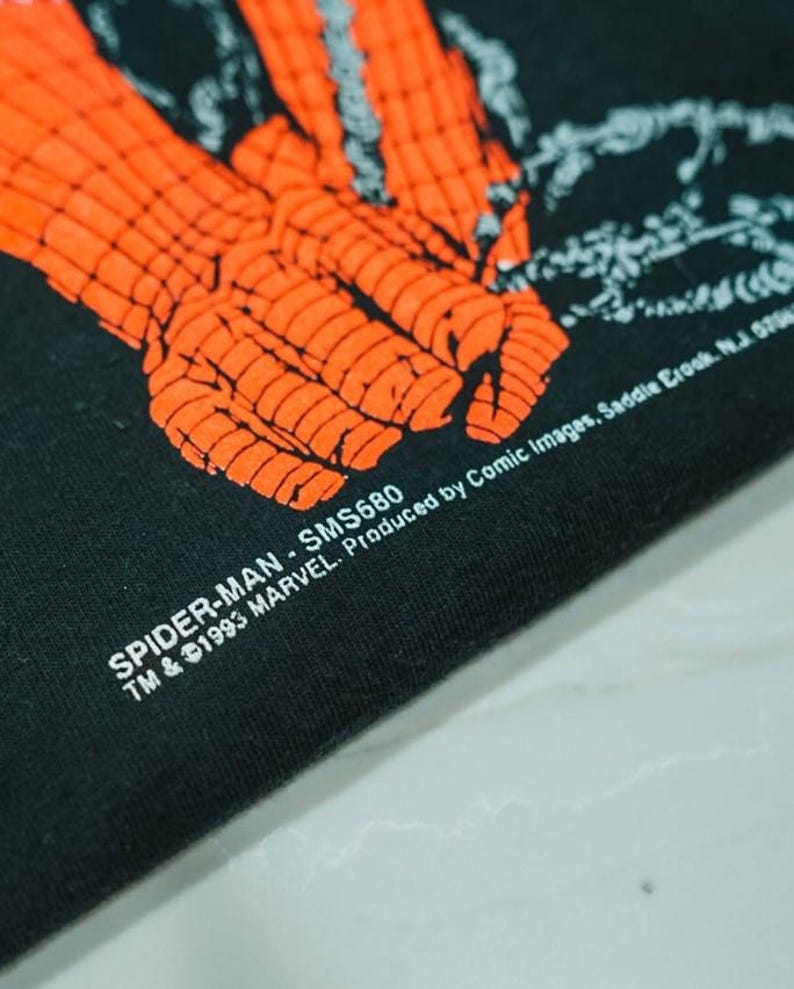 May include: A black fabric garment featuring a bright orange graphic of a gloved hand with a webbed pattern. The text reads: SPIDER-MAN - SMS680 TM & &copy;1993 MARVEL. Produced by Comic Images, Saddle Brook, N.J. 07662.