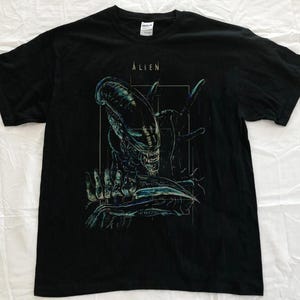 May include: Black t-shirt featuring a detailed graphic of an Alien creature with the word "ALIEN" above the image. The design is in shades of blue, green, and white.