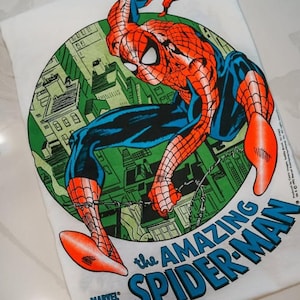 May include: A white t-shirt featuring a circular graphic of Spider-Man in a red and blue suit crouching over a green city skyline. The text reads The Amazing Spider-Man in blue letters, with a small Marvel Comics logo in the bottom left corner.