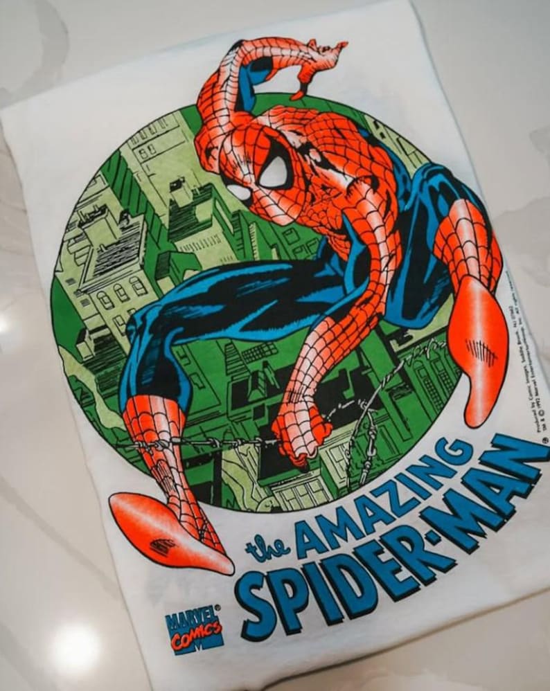 May include: A white t-shirt featuring a circular graphic of Spider-Man in a red and blue suit crouching over a green city skyline. The text reads The Amazing Spider-Man in blue letters, with a small Marvel Comics logo in the bottom left corner.