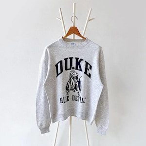 May include: A heather gray sweatshirt with the word "DUKE" in navy blue arched across the chest. Below the word "DUKE" is a graphic of a figure in a cape holding a sword. The words "BLUE DEVILS" are printed below the graphic in navy blue.