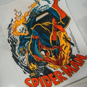 May include: A white t-shirt featuring a graphic illustration of Spider-Man and Ghost Rider in a dynamic pose surrounded by orange flames and debris. The text SPIDER-MAN is printed in large red and yellow letters at the bottom right corner.