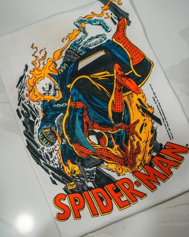May include: A white t-shirt featuring a graphic illustration of Spider-Man and Ghost Rider in a dynamic pose surrounded by orange flames and debris. The text SPIDER-MAN is printed in large red and yellow letters at the bottom right corner.