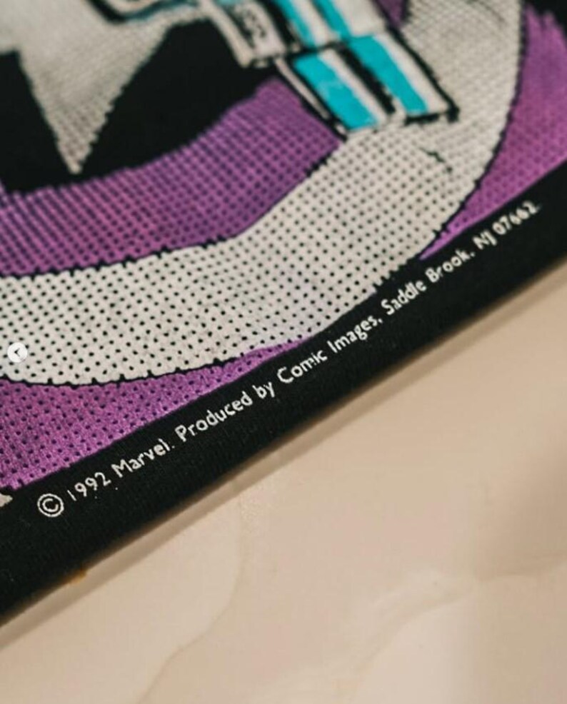 May include: A black fabric item featuring a purple and white graphic design with a halftone dot pattern. The text printed on the fabric reads: 1992 Marvel. Produced by Comic Images, Saddle Brook, NJ 07662. The material has a visible mesh texture.