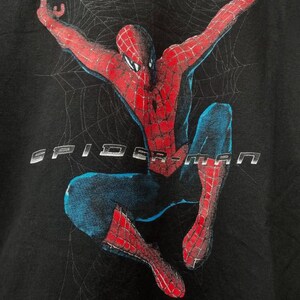 May include: A black t-shirt featuring a graphic print of the character Spider-Man in a dynamic pose with arms raised against a faint spiderweb background. The text SPIDER-MAN is printed in a metallic silver font across the center of the chest.