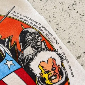 May include: A white fabric graphic print featuring a 1990 Marvel Entertainment Group copyright. The design includes a stylized character with a yellow face and dark hair, a black helmeted figure, and a red, white, and blue shield graphic.
