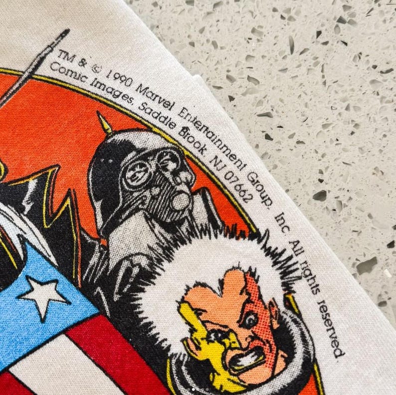 May include: A white fabric graphic print featuring a 1990 Marvel Entertainment Group copyright. The design includes a stylized character with a yellow face and dark hair, a black helmeted figure, and a red, white, and blue shield graphic.