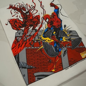 May include: A white t-shirt featuring a graphic illustration of Spider-Man and Carnage in a battle scene. The print shows Spider-Man in red and blue leaping over a brick wall while Carnage in red stands above. Text reads MARVEL COMICS and SPIDEY/CARNAGE.