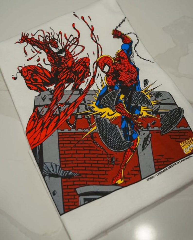 May include: A white t-shirt featuring a graphic illustration of Spider-Man and Carnage in a battle scene. The print shows Spider-Man in red and blue leaping over a brick wall while Carnage in red stands above. Text reads MARVEL COMICS and SPIDEY/CARNAGE.