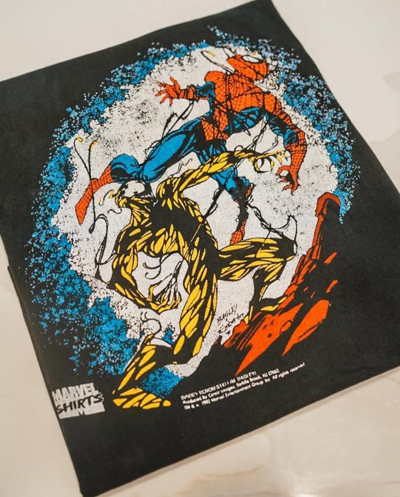 May include: A black t-shirt featuring a graphic print of Spider-Man in red and blue fighting a yellow and black creature. The design includes a blue speckled circular background, the Marvel Shirts logo, and copyright text dated 1992 at the bottom.