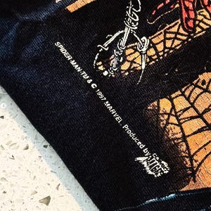 May include: A black fabric garment featuring a graphic print with orange and black spiderweb patterns. The text reads SPIDER-MAN TM & &copy; 1997 MARVEL. Produced by Giant. The print shows a portion of a character illustration with detailed line work.