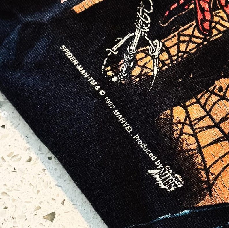 May include: A black fabric garment featuring a graphic print with orange and black spiderweb patterns. The text reads SPIDER-MAN TM & &copy; 1997 MARVEL. Produced by Giant. The print shows a portion of a character illustration with detailed line work.
