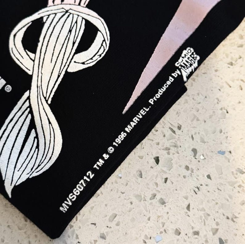 May include: A black fabric item featuring a white graphic print of a stylized circular element and text that reads MVS60712 TM & &copy; 1996 MARVEL. Produced by COMIC IMAGES. The design includes a white curved shape and a portion of a larger graphic.