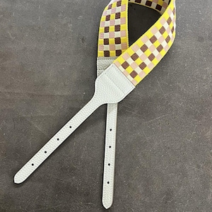 May include: A patterned shoulder strap with a woven design in yellow, brown, and pink squares. The strap has white leather ends with adjustment holes, designed for a bag or purse.