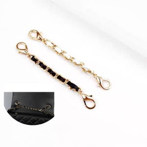 May include: Two gold-tone metal chain bag extenders, one woven with black leather-like material and one with white leather-like material, each featuring a lobster claw clasp at both ends, shown alongside a black quilted handbag with a chain strap.