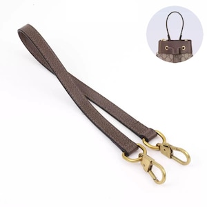 May include: A brown leather handbag strap with gold-coloured metal clasps and rings. The strap is designed to attach to a small handbag, as shown in the inset image. The leather has a textured appearance.