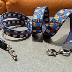 May include: A blue, red, and yellow checkered pattern strap with silver-tone metal clasps. The strap is made of woven fabric and has a leather end piece. The strap is designed for a bag or purse.
