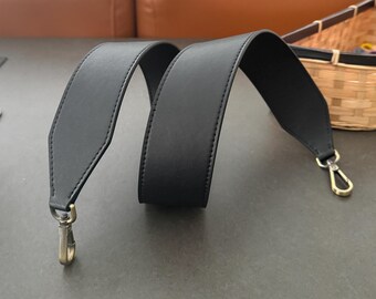 May include: A black leather bag strap with gold-colored metal clasps. The strap is wide and has a stitched edge. The clasps are attached to the ends of the strap, ready to be attached to a bag.