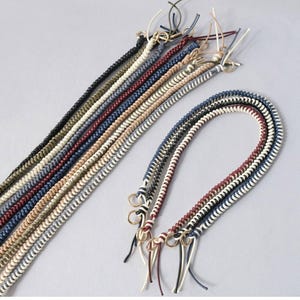 May include: Braided cord straps in various colors, including black, navy, red, and cream. Each strap features a gold-colored ring and multiple strands. These straps are suitable for bags or other accessories.