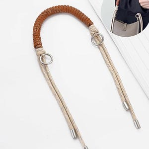 Könnte beinhalten: Ein brauner und beiger Handtaschengriff mit einem geflochtenen Oberteil und seilartigen Riemen. Der Griff verfügt über silberfarbene Metallringe und Endkappen. Das Design ist für verschiedene Taschenstile geeignet.