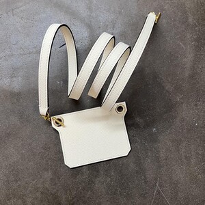以下が含まれることがあります： 白革のIDホルダー。ストラップとゴールドトーンの金具付き。ホルダーは長方形で、表面はテクスチャ加工、黒いトリム。ストラップはループ状で、ゴールドの留め金とグロメットで取り付けられています。