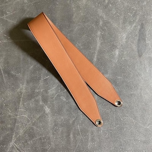 Könnte beinhalten: Ein hellbrauner Leder-Taschenriemen mit silberfarbenen Metallösen. Der Riemen ist hellbraun und scheint neu zu sein. Der Riemen ist für die Befestigung an einer Tasche vorgesehen.