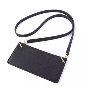 May include: Black crossbody phone bag with a long strap. The rectangular bag has gold-colored accents and a textured surface. The strap is thin and appears to be made of the same material as the bag. The bag is designed to be worn across the body.