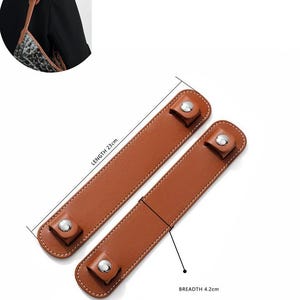 May include: Two brown leather bag handles with silver snap closures. The handles are 23 cm long and 4.2 cm wide, with white stitching along the edges. The handles are shown on a white background.