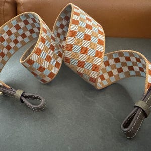 May include: A woven strap with a checkered pattern in shades of brown, orange, and blue. The strap has dark brown leather ends with a loop and a gold-colored rivet. The strap is designed for a bag or purse.