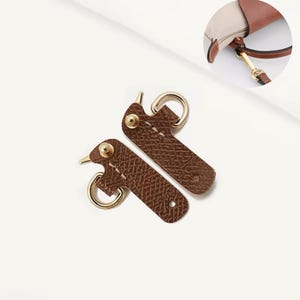 May include: Two brown leather bag strap connectors with gold-colored hardware. Each connector has a D-ring and is secured with a gold rivet. The connectors are designed to attach to a bag strap.