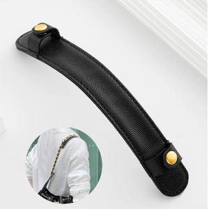 May include: A black leather bag handle with gold-colored button closures. The handle is curved and textured, designed to be attached to a handbag. The image includes a circular inset showing the handle in use on a bag.