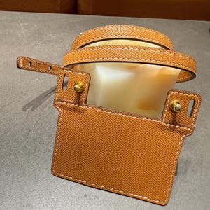 May include: A tan leather handbag with a rectangular front flap and a curved top handle. The bag features gold-colored hardware and white stitching. The interior is a light cream color.