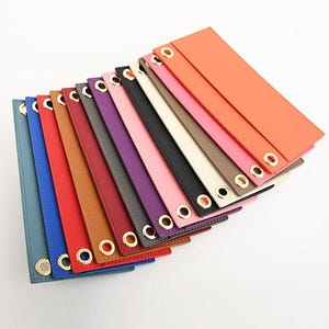 May include: A collection of colorful leather straps, each with a gold-toned grommet at the end. The straps are arranged in a fan shape, showcasing a variety of colors including blue, red, brown, purple, pink, and orange. The leather appears smooth and the grommets are evenly spaced.
