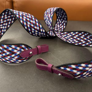 Puede incluir: Una correa tejida con un patrón geométrico en rojo, blanco y azul. La correa tiene bordes negros y se combina con dos extremos de cuero púrpura con herrajes plateados. Diseñada para un bolso o cartera.