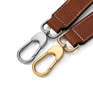 May include: Close-up of two metal clasp hooks with brown leather straps. One clasp is silver, the other is gold. The leather straps are brown with white stitching. The clasps are designed to attach to a bag or other accessory.
